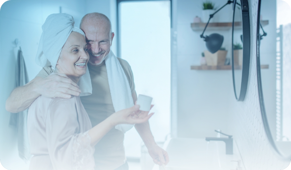 Happy senior couple in their bathroom. The man has his arm around the woman, who is holding a jar of moisturizing skin care.